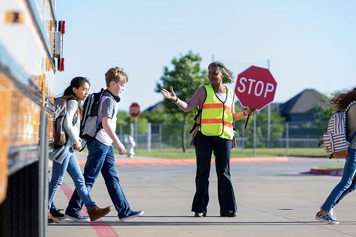 As students cross the street, a senior female crossing guard holds up her stop sign to traffic and protects the students. She waves to the students to keep walking.