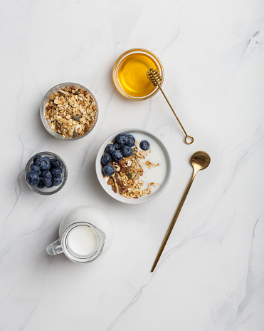Porridge with nuts, yogurt, blueberries, honey with honey stick, with golden tableware on white marble background. Copy space for text, flat lay