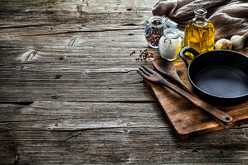 Cooking backgrounds: Top view of a black cast iron pan, salt, olive oil, peppercorns arranged at the right of a rustic wooden kitchen table leaving a useful copy space for text and/or logo at the center left. A brown tablecloth is at the top right corner of the table. Predominant color is brown. Low key DSRL indoors photo taken with Canon EOS 5D Mk II and Canon EF 24-105mm f/4L IS USM Wide Angle Zoom Lens