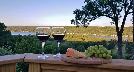 2 glasses filled with red wine, a plate of cheddar cheese wedges and green grapes with a beautiful lake/water panoramic view nearing the end of a long day; wine tasting, relaxing, enjoyment. wine and cheese concepts.