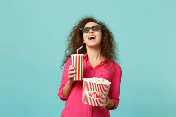 Laughing african girl in 3d imax glasses watching movie film hold popcorn cup of soda isolated on blue turquoise background in studio. People emotions in cinema, lifestyle concept. Mock up copy space
