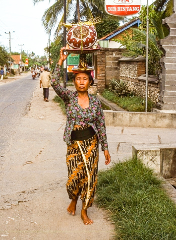 Nostalgia. Bali Indonesia, Kuta Beach 1976 Traditionally dressed balinese woman with offerings on her head is walking on the street, Houses, people and restaurant with a sign advertising beer in the background.