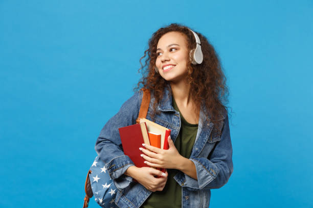 joven chica afroamericana estudiante adolescente en ropa de mezclilla, auriculares de mochila aislados en el retrato de fondo de pared azul de estudio. educación en el concepto universitario universitario de secundaria. mock up copia espacio - child lifestyles isolated blue fotografías e imágenes de stock