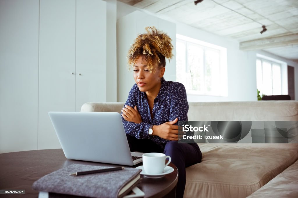 Confident businesswoman looking at laptop Confident businesswoman looking at laptop. Female financial expert is sitting on sofa in office. She is having curly hair. Corporate Business Stock Photo