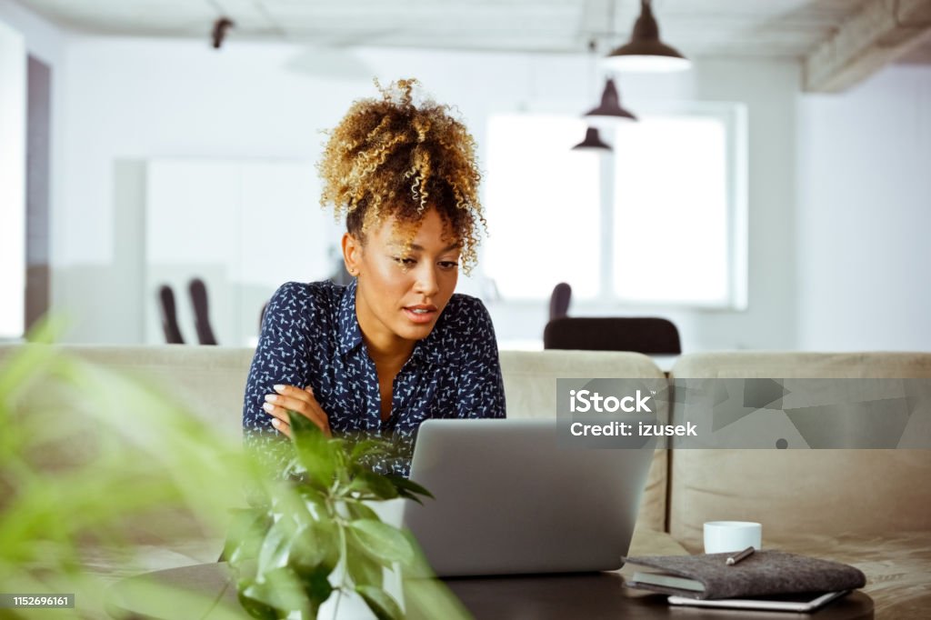 Confident financial advisor looking at laptop Confident financial advisor looking at laptop. Corporate professional is concentrated towards her work. She is having curly hair in office. UK Stock Photo