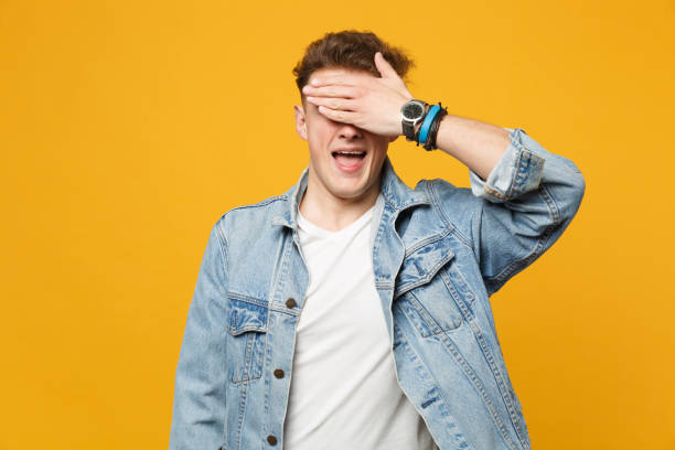 Portrait of funny young man in denim casual clothes keeping mouth open covering eyes with hand isolated on yellow orange wall background. People sincere emotions lifestyle concept. Mock up copy space. Portrait of funny young man in denim casual clothes keeping mouth open covering eyes with hand isolated on yellow orange wall background. People sincere emotions lifestyle concept. Mock up copy space hair covering eyes stock pictures, royalty-free photos & images
