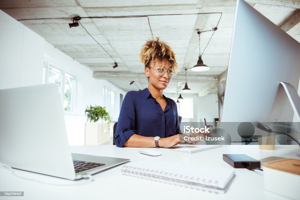 Confident businesswoman using computer at desk Confident businesswoman using computer at desk. Female financial expert is sitting in office. She is having curly hair. Computer Monitor Stock Photo