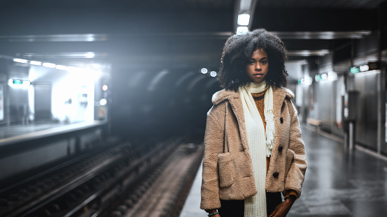 A dazzling African-American girl with curly lush Afro hair in a demo-season coat and scarf is standing on the subway platform waiting for the train; cute biracial female in the metro