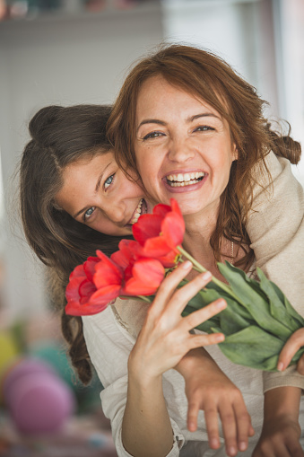 Smiling mid adult woman cuddling her teenage daughter while holding a bouquet of beautiful red tulips and looking at camera.