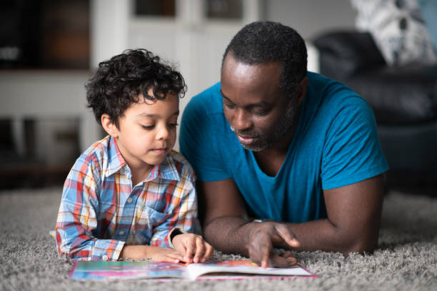 Father reading to son A young father of African descent is lying on the carpet in their living room and reading to his son. parent and child reading stock pictures, royalty-free photos & images