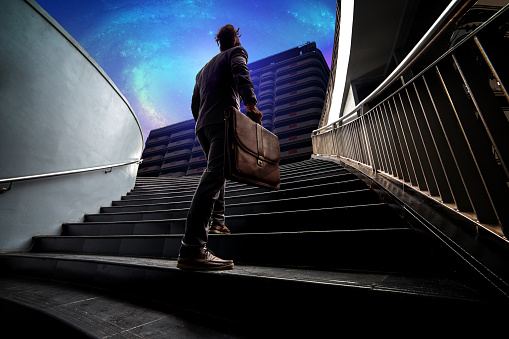The young man in a formal suit holds a traditional leather bag, walking up the stairs to the future city and galaxy. He have ambition for step up to next generation.