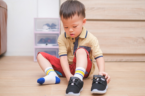 Little Asian 2 - 3 years old toddler boy sitting and concentrate on putting on his black shoes / sneakers, Get toddler out the door, Encourage Self-Help Skills in Children, Develop Confidence concept