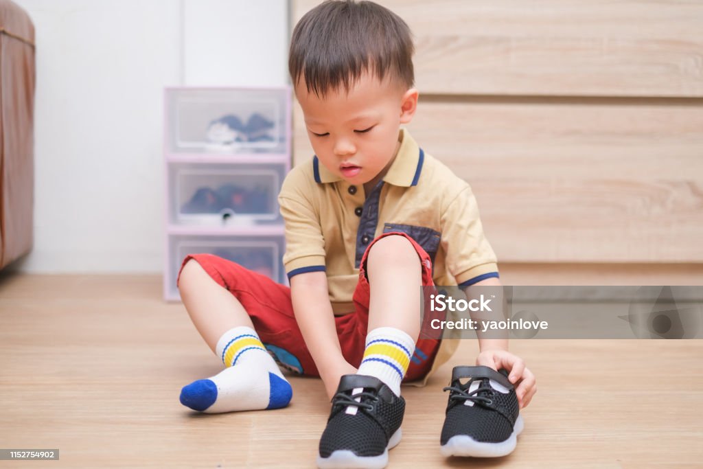 Asian 2 - 3 years old toddler boy sitting and concentrate on putting on his black shoes / sneakers Little Asian 2 - 3 years old toddler boy sitting and concentrate on putting on his black shoes / sneakers, Get toddler out the door, Encourage Self-Help Skills in Children, Develop Confidence concept Child Stock Photo