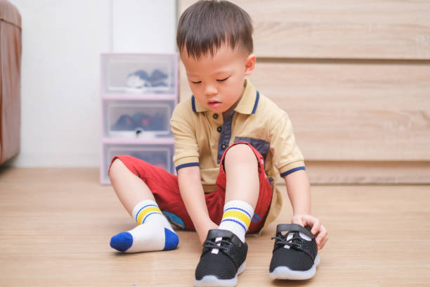 Asian 2 - 3 years old toddler boy sitting and concentrate on putting on his black shoes / sneakers stock photo