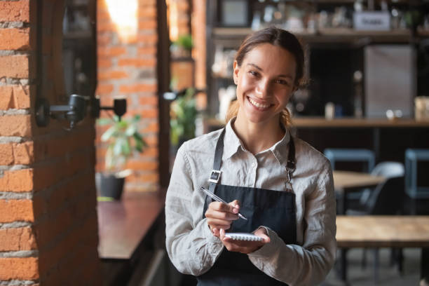 Head shot portrait of smiling waitress ready to take customer order Head shot portrait of smiling waitress wearing black apron ready to take customer order, attractive woman with notebook and pen in hands looking at camera, standing in cozy coffeehouse, good service cafe racer stock pictures, royalty-free photos & images
