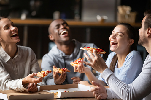 Diverse friends eating pizza, happy colleagues or students having fun together in cafe, men and women laughing at funny joke, holding Italian junk food slices in hands at meeting in cafeteria