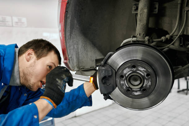 mecânico masculino com a boca aberta que desgasta luvas pretas e o uniforme azul, prendendo a lanterna elétrica e examinando com cuidado pneumáticos ou pastilhas de freio do carro levantado na auto oficina. conceito do serviço e do técnico do carro - lona de freio - fotografias e filmes do acervo