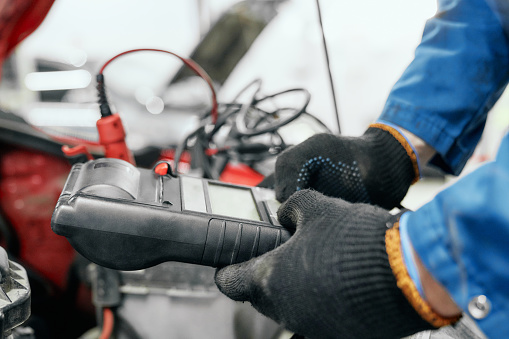 Close up of black tester digital multimeter with lot of wires for checking charge of accumulator that holding mechanic's hands in dirty gloves. Professional man doing diagnostic for old car in service