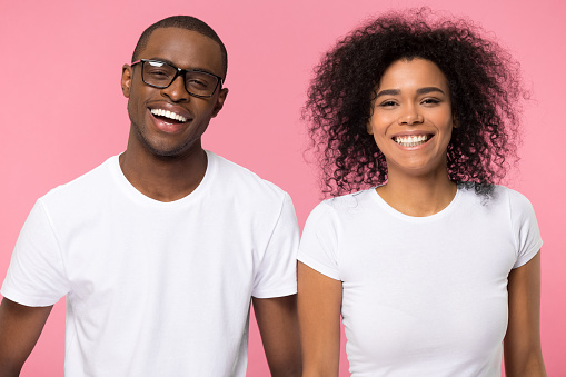 Head shot portrait african couple in white t-shirts on pink blank laughing looking at camera, guy in glasses girl afro curly hair people feel happy toothy smile positive face expressions concept image