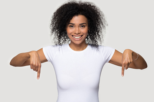 Attractive mixed race woman in white t-shirt curly afro hairs pointing fingers down looking at camera standing over grey blank wall advertise product beauty salon professional care copy space for text