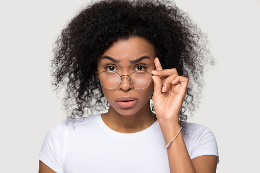 Studio portrait of shocked african young woman teacher wearing white t-shirt lowering glasses looking at camera feels confused surprised received amazing stunned news posing isolate on grey background