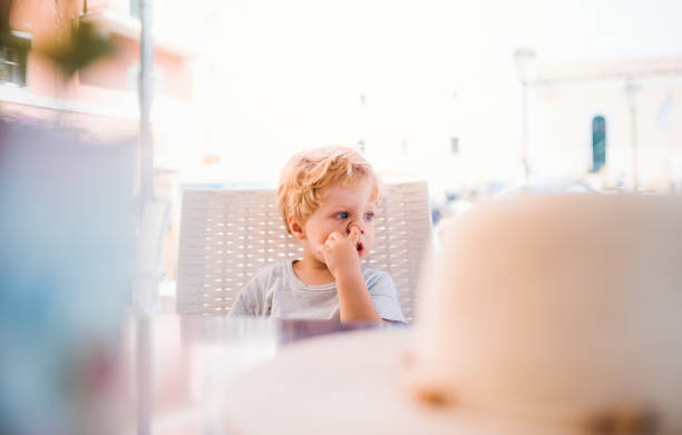 mały chłopiec siedzi w restauracji na świeżym powietrzu na wakacjach, zbierając nos. - picking nose zdjęcia i obrazy z banku zdjęć