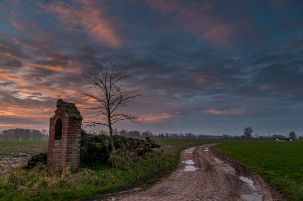 sunrise over a country road. Impression of a worn-down memorial in the flemish country side, right next to an unpaved country road, flooded with pink light from an early morning sunrise. julian california stock pictures, royalty-free photos & images