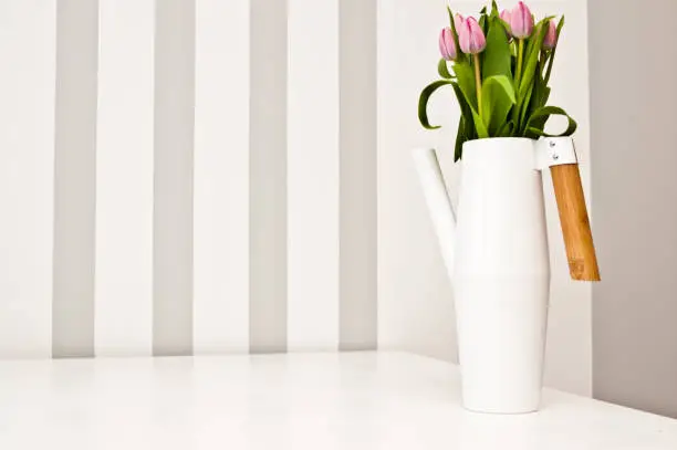 A white modern kettle shaped flower vase on the kitchen table with fresh tulip flowers.