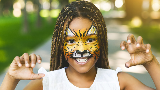 Funny little tiger. Adorable african-american girl with creative face painting roaring, playing wild cat outdoors