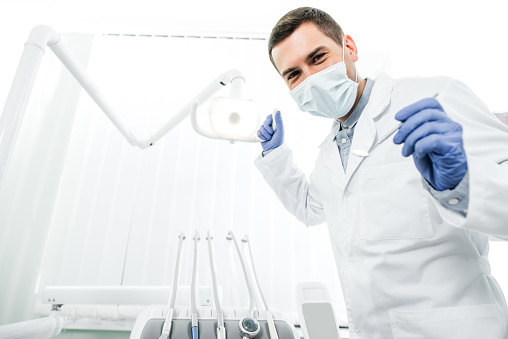 dentist in latex gloves and mask holding dental instrument and dental lamp
