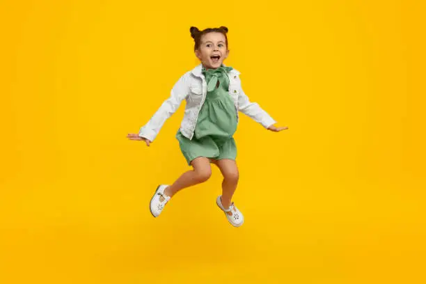 Adorable little girl in stylish dress and jacket yelling and leaping up in excitement against bright yellow background