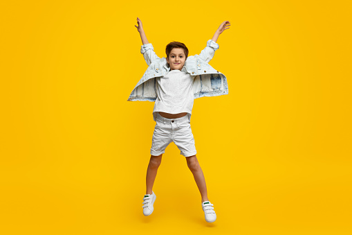 Full length stylish youngster with raised hands looking at camera and leaping up against yellow background