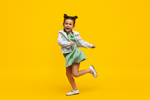 Adorable little girl in trendy dress and jacket cheerfully smiling and twisting on one leg while dancing against bright yellow background