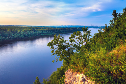 A large cliff overgrown with green grass and trees dominate over the White River in the evening at dusk during sunset under a clear blue sky.