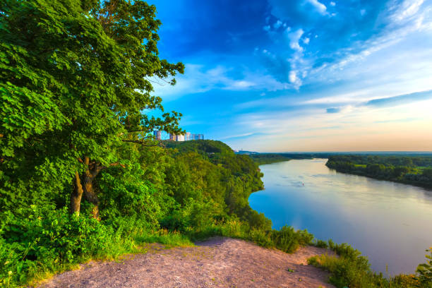 A large cliff overgrown with green grass and trees dominate over the White River in the evening at dusk during sunset under a clear blue sky.