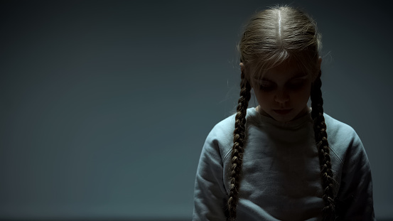 Depressed female kid looking down, standing alone in dark room, loneliness