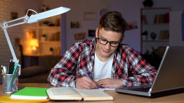 istock Student in glasses doing homework, writing essay, open book and laptop on table 1152831310
