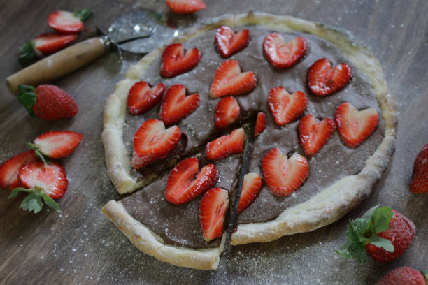 Image of homemade chocolate pizza with strawberries and icing sugar, baked pizza crust filled with hazelnut chocolate spread and topped with romantic red love heart shape slices of strawberries / strawberries fruit, ready to be sliced and eaten as pudding Stock photo of strawberry and chocolate pizza, romantic strawberries hearts with chocolate served for Valentine's Dat dinner, healthy eating breakfast five a day fruit and vegetables. pizza topping stock pictures, royalty-free photos & images