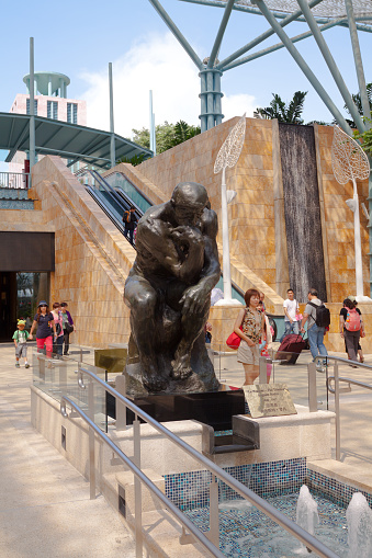 Asian tourists are passing copy of The Thinker on Sentosa Island, scene on steps on way up to Merlion copy statue. Square area between buildings and hotels and casino on island.