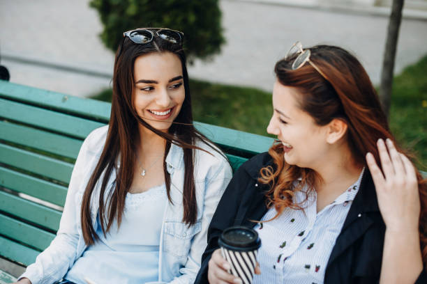 istock Two young caucasian female talking smiling while sitting on a bench outside drinking coffee. 1152839111