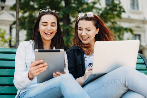 istock Two lovely woman laughing while looking at a tablet screen while one is holding a laptop on the legs sitting on bench outside. 1152839154