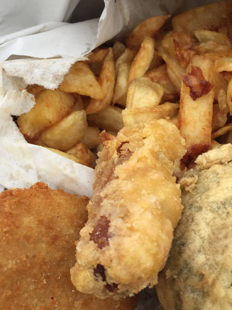 Stock photo of eating takeaway fish, battered sausage and chips, fishcake, mushy pea fritter British chippy shop in a cardboard box with salt and vinegar, eating fish and chips at the beach. imagen de un pez grasoso de cerca, salchichas y papas fritas, torta de pescado frito en pan rallado, buñuelo de guisante en caja de cartón y papel blanco con sal y vinagre de temporada, mariscos para llevar alimentos poco saludables, salchichas maltratad