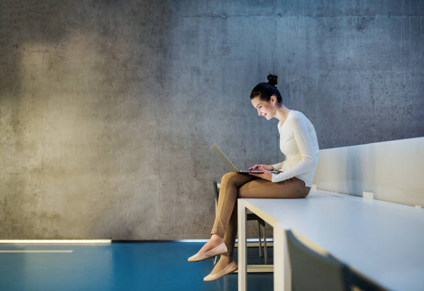 Young student or businesswoman sitting on desk in room in a library or office, using laptop. A portrait of a young student or businesswoman sitting on desk in room in a library or office, using laptop. woman legs open stock pictures, royalty-free photos & images