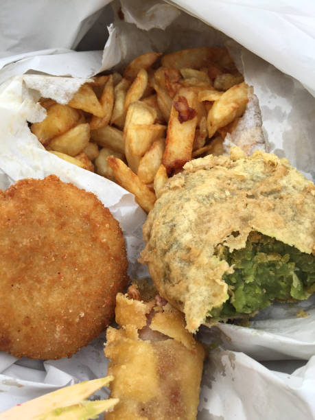 Stock photo of eating takeaway fish, battered sausage and chips, fishcake, mushy pea fritter British chippy shop in a cardboard box with salt and vinegar, eating fish and chips at the beach. imagen de un pez grasoso de cerca, salchichas y papas fritas, torta de pescado frito en pan rallado, buñuelo de guisante en caja de cartón y papel blanco con sal y vinagre de temporada, mariscos para llevar alimentos poco saludables, salchichas maltratad