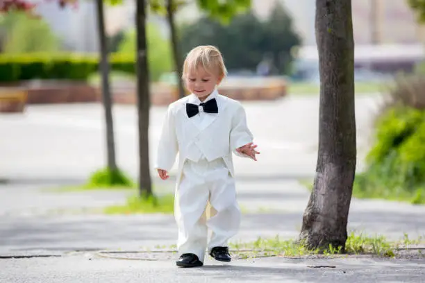 Photo of Beautiful toddler boy in tuxedo, playing in a park on a wedding day