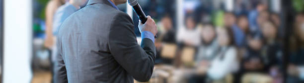 Focus on microphone held by panel speaker on stage during presentation. Executive manager presenter at corporate conference talking to audience.  Business leadership CEO lecture during seminar. stock photo