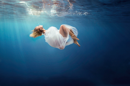 View under water of a fantasy girl wrapped in fine white cloth, sank in blue deep water of ocean, against dark sea background with bright sunshine. Horizontal view.