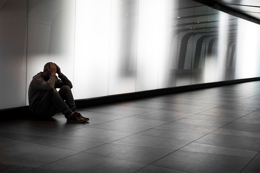 Color image depicting a side view of a young homeless man sitting alone in a futuristic illuminated subway tunnel in the city. The man is clearly sad, and has an unkempt beard. He has his head in his hands in a gesture of despair. He sits with his back to the wall of the tunnel while the diminishing perspective of the subway tunnel recedes into the distance. People are defocused in the distance as they walk past, ignoring the young homeless man. Room for copy space.