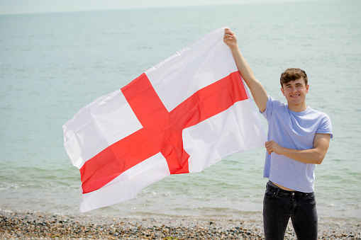 Young adult Caucasian male holding on a beach holding the flag of England