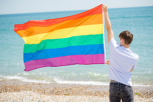 Young adult Caucasian male holding on a beach holding the Pride flag